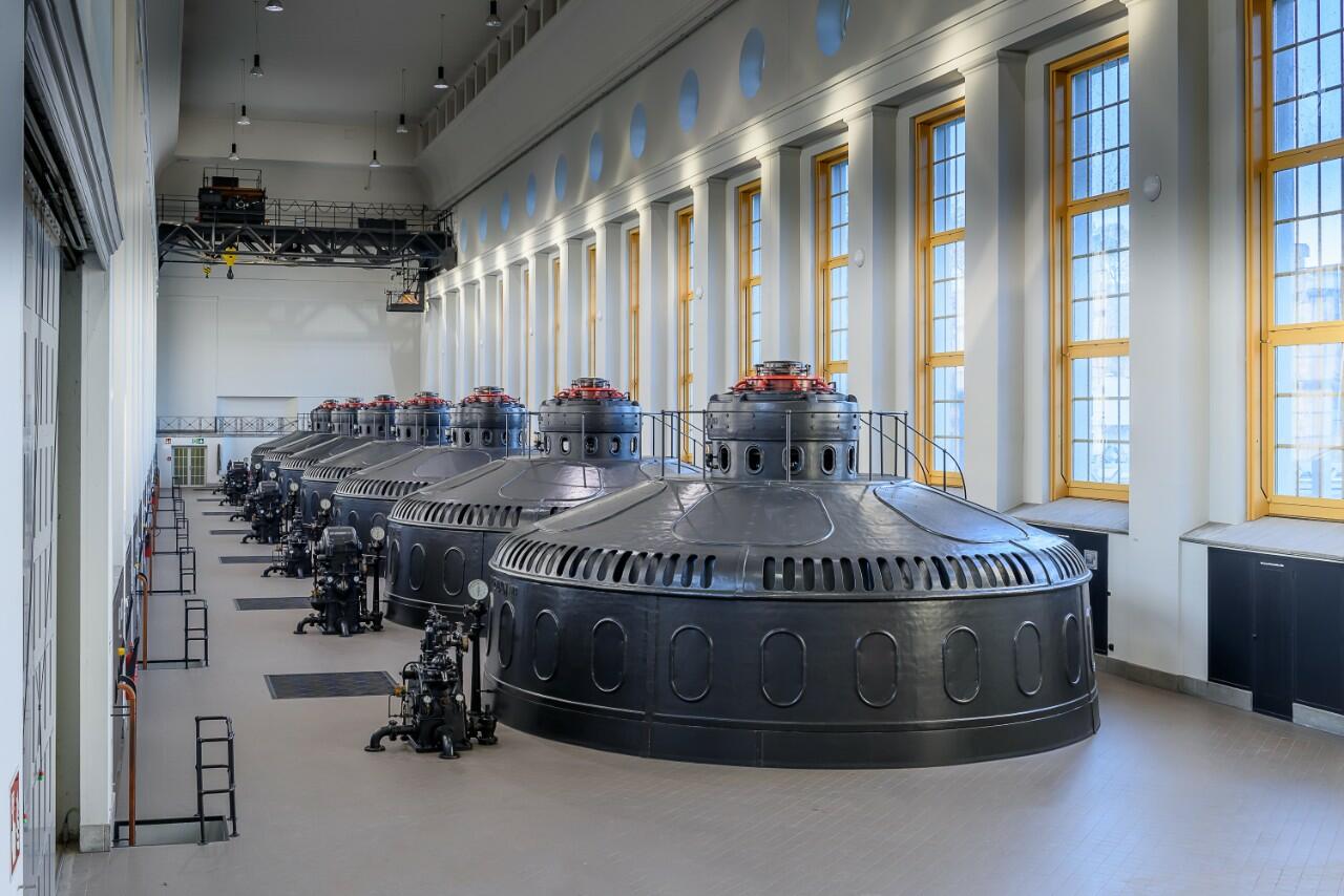 Maschinenhalle eines Wasserkraftwerks (Eglisau) mit einer Reihe grosser, schwarzer Generatoren/Turbinengehäuse. Rechts fallen Sonnenstrahlen durch hohe Fenster mit gelben Rahmen auf den Boden und die Anlagen.
